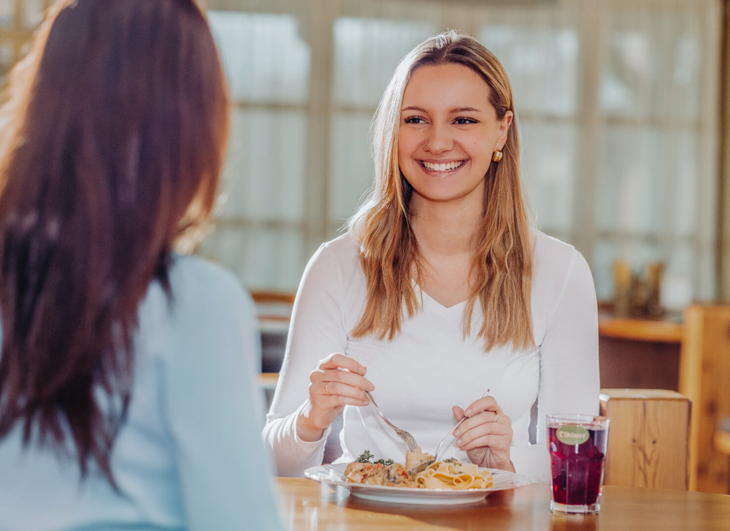 Zwei Frauen unterhalten sich beim Essen. Eine Frau ist im Fokus, lächelt und genießt ein Nudelgericht.
